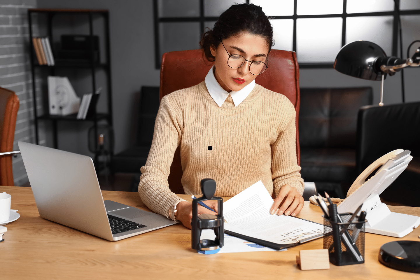 A professional woman reviewing documents at her desk beside a laptop and office supplies.