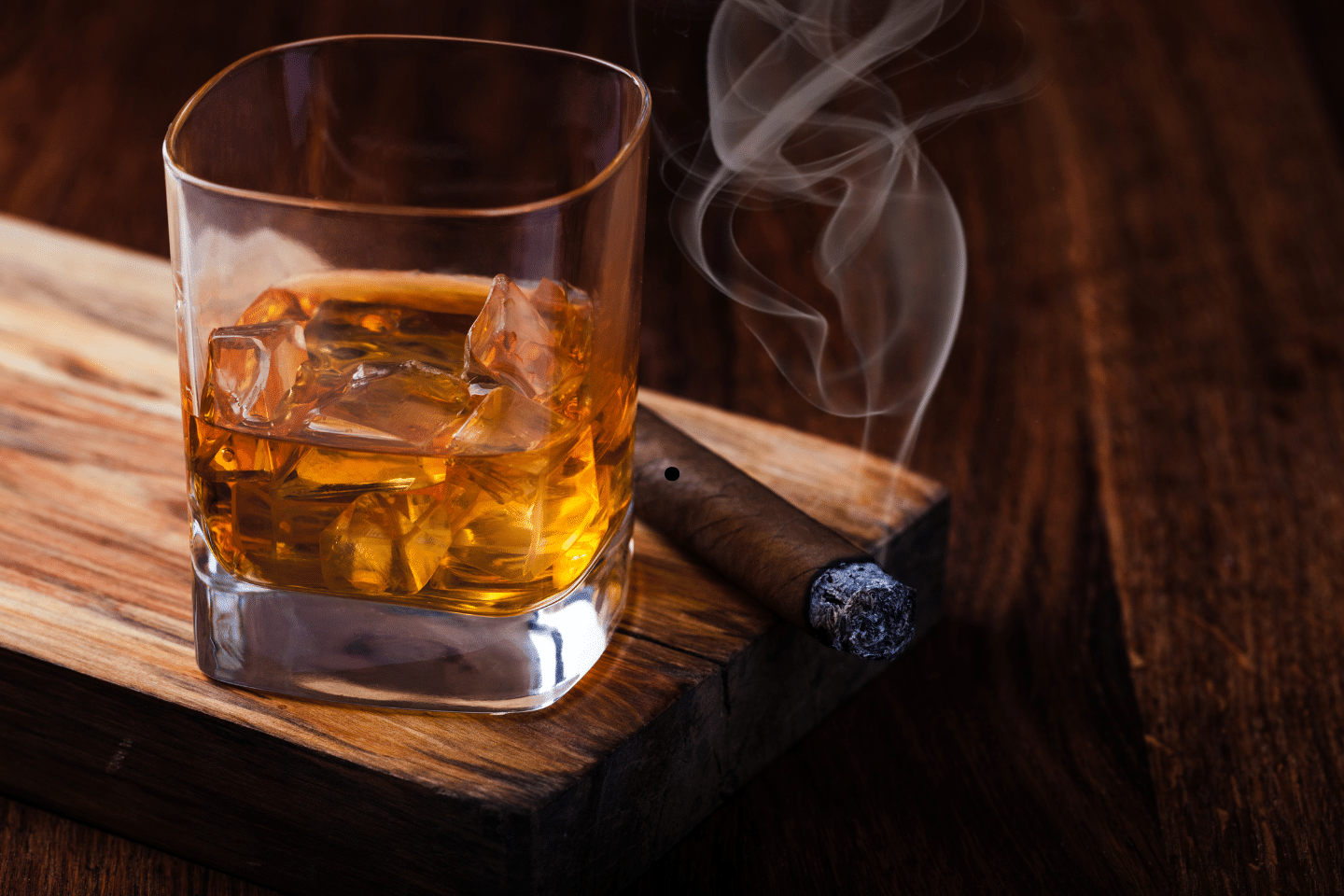 A glass of whiskey with ice sits beside a smoldering cigar on a wooden board.