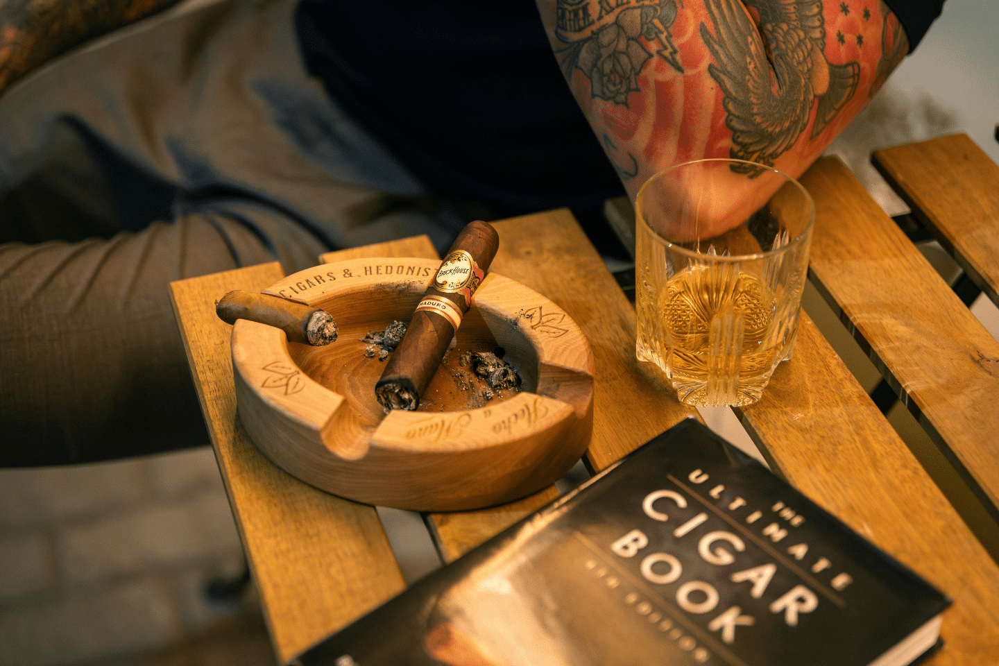 A tattooed person enjoys whiskey and a cigar next to a wooden ashtray and cigar book.