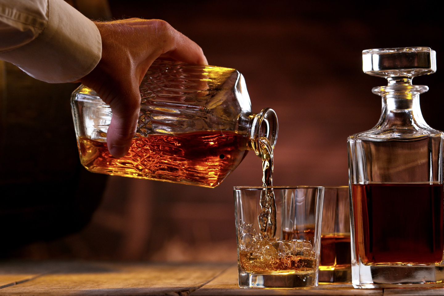 Hand pouring whiskey from a decanter into a glass on a wooden surface.