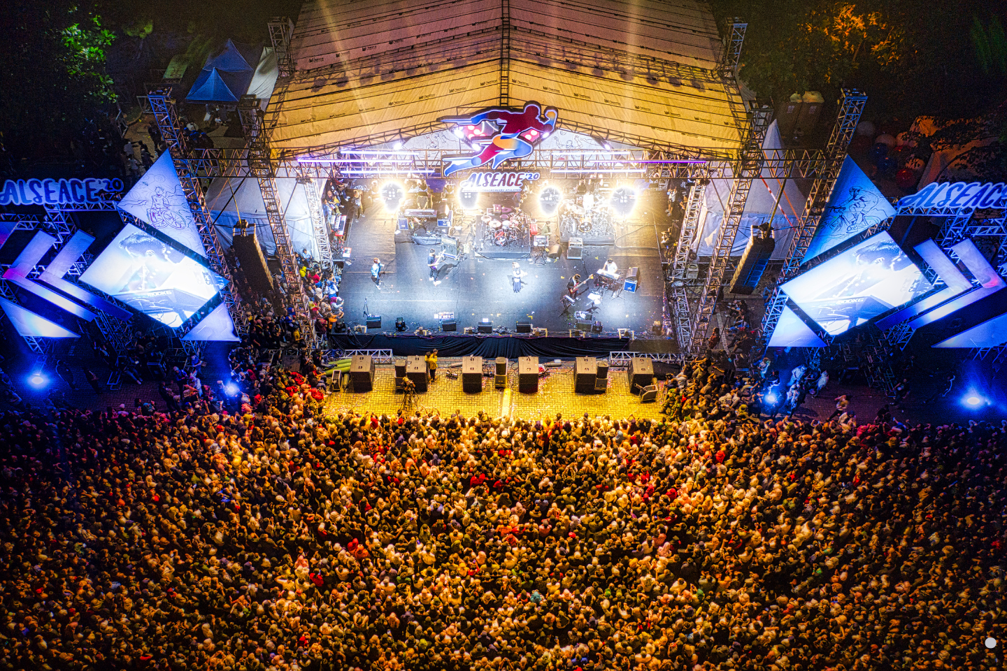 A large illuminated festival stage viewed from above, surrounded by a packed crowd.