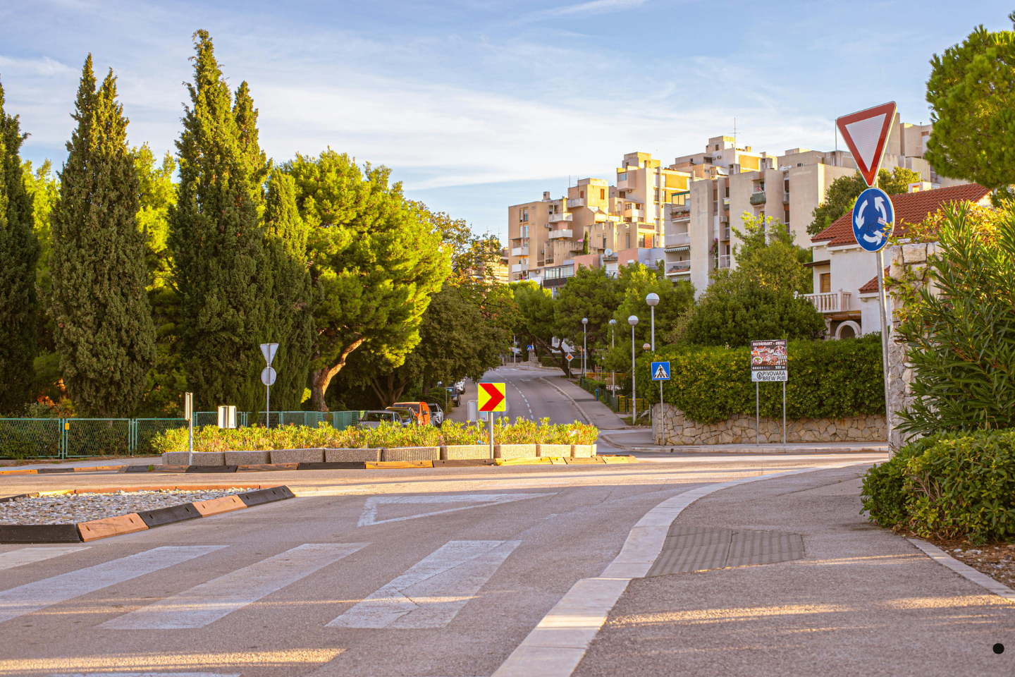 A quiet urban street lined with trees, road signs, and residential buildings in warm daylight.
