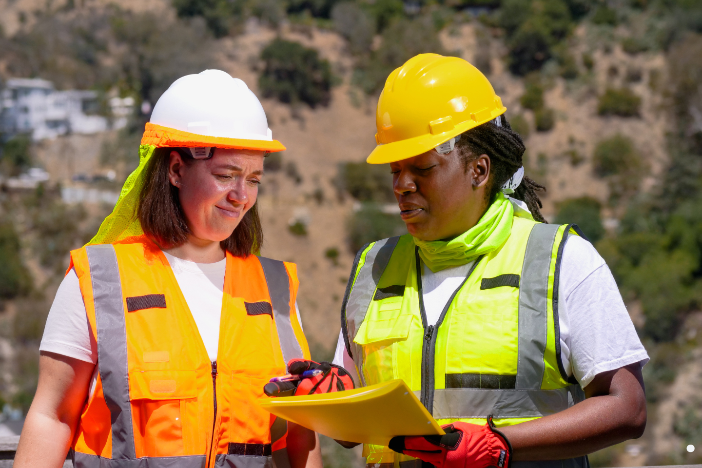 Two construction workers reviewing documents outdoors while wearing safety vests and helmets.