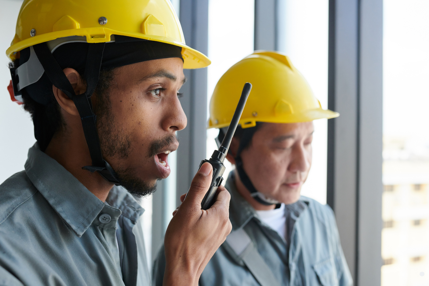 Two construction workers in yellow helmets communicating with a walkie-talkie on-site.