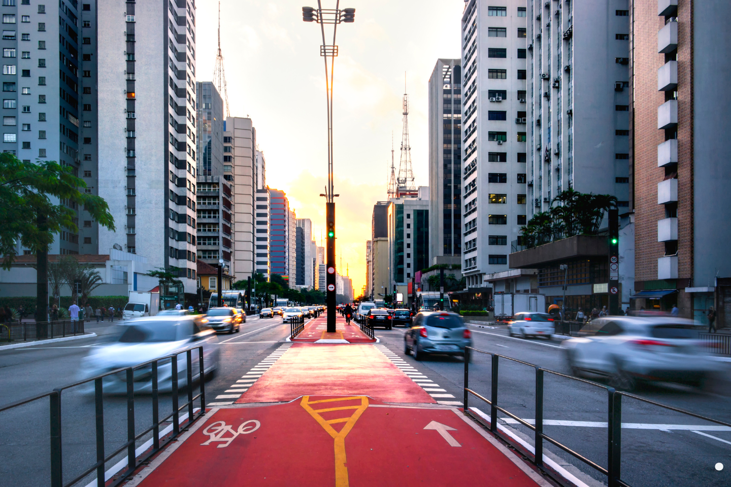 A busy city boulevard at sunset with blurred car movement and a red bike lane running through tall skyscrapers.