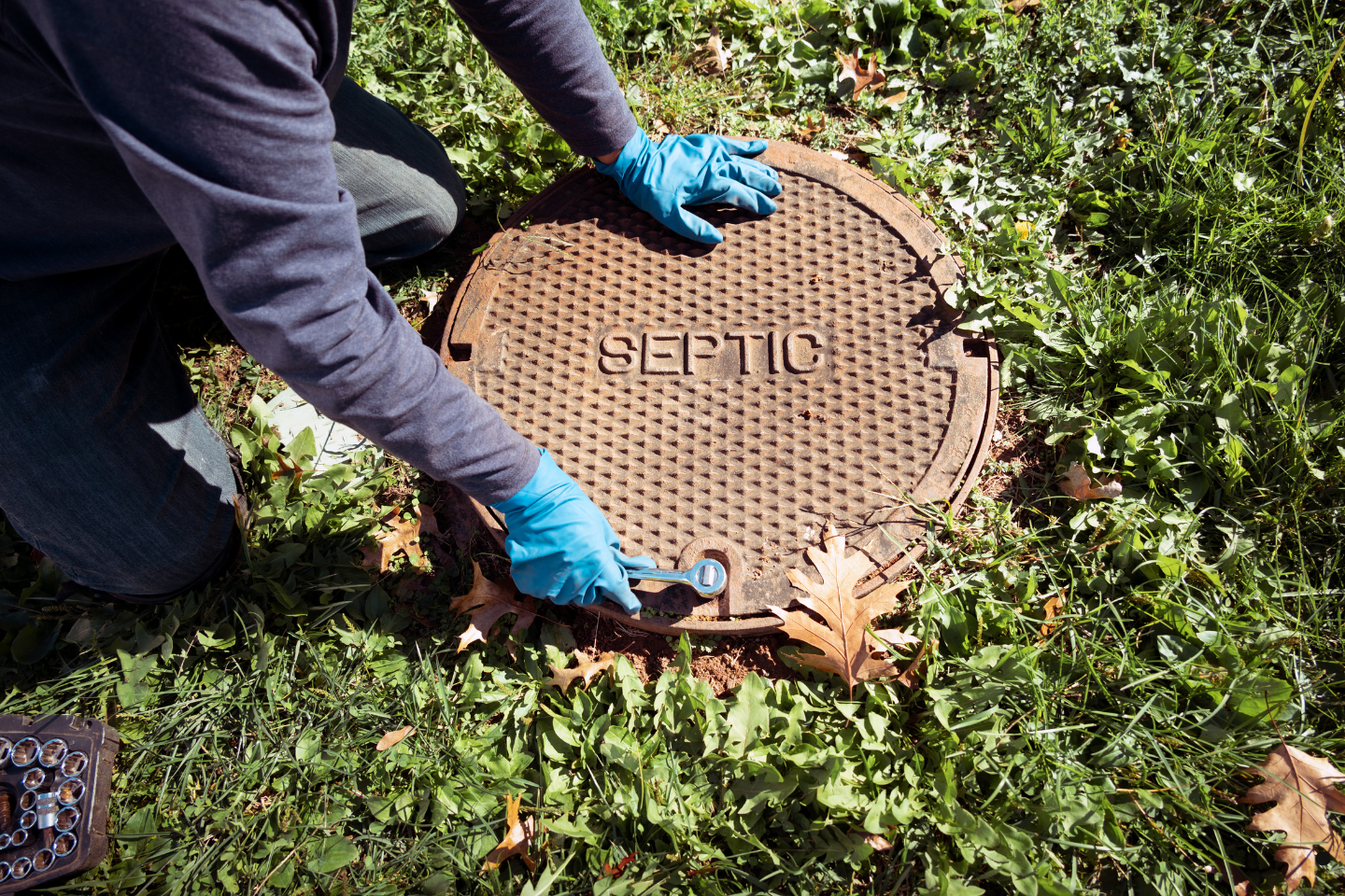 Technician wearing gloves opening a septic tank cover in a grassy yard.