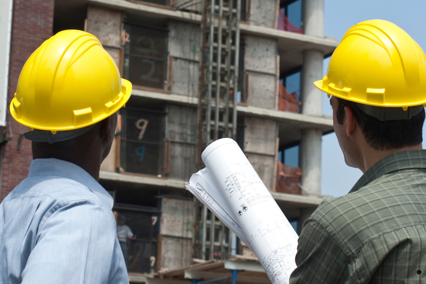 Two workers in yellow hard hats review building plans in front of a multi-story construction project.