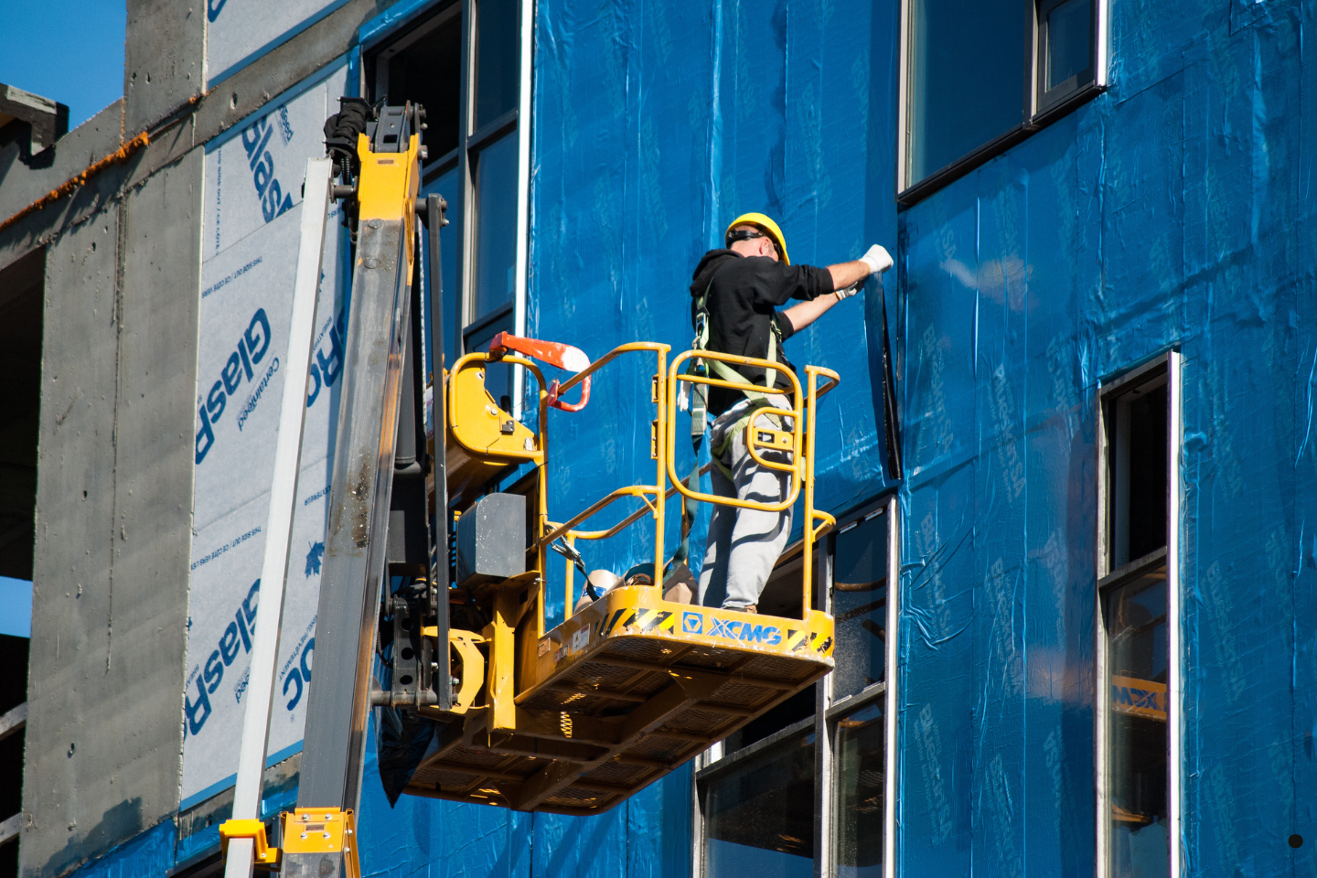 A construction worker on a lift installs or secures blue sheathing on a building exterior.