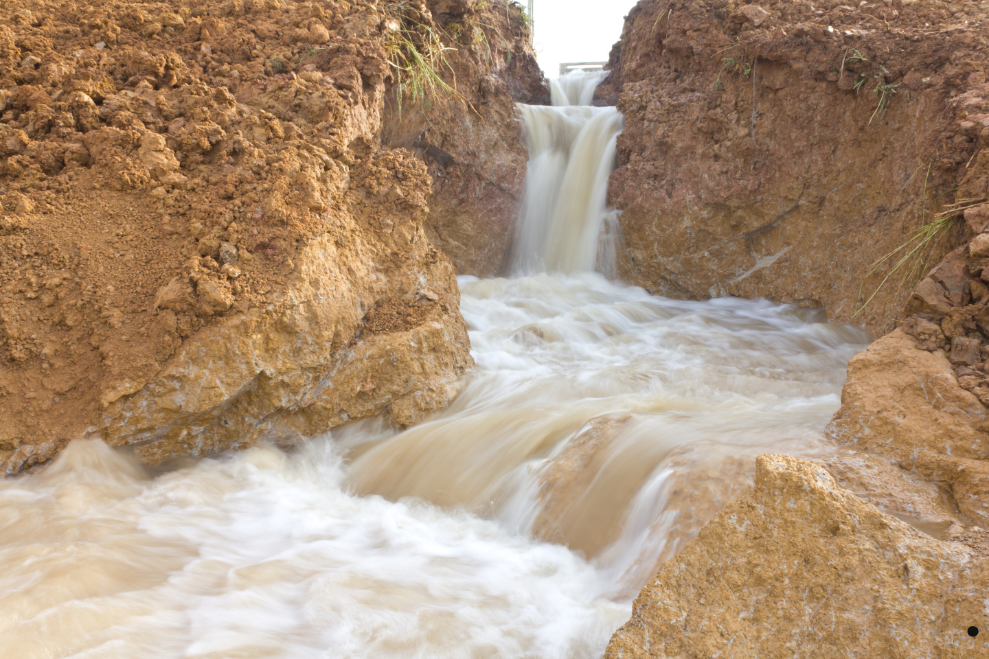 Rapid stormwater runoff flowing through an eroded dirt channel resembling a small waterfall.