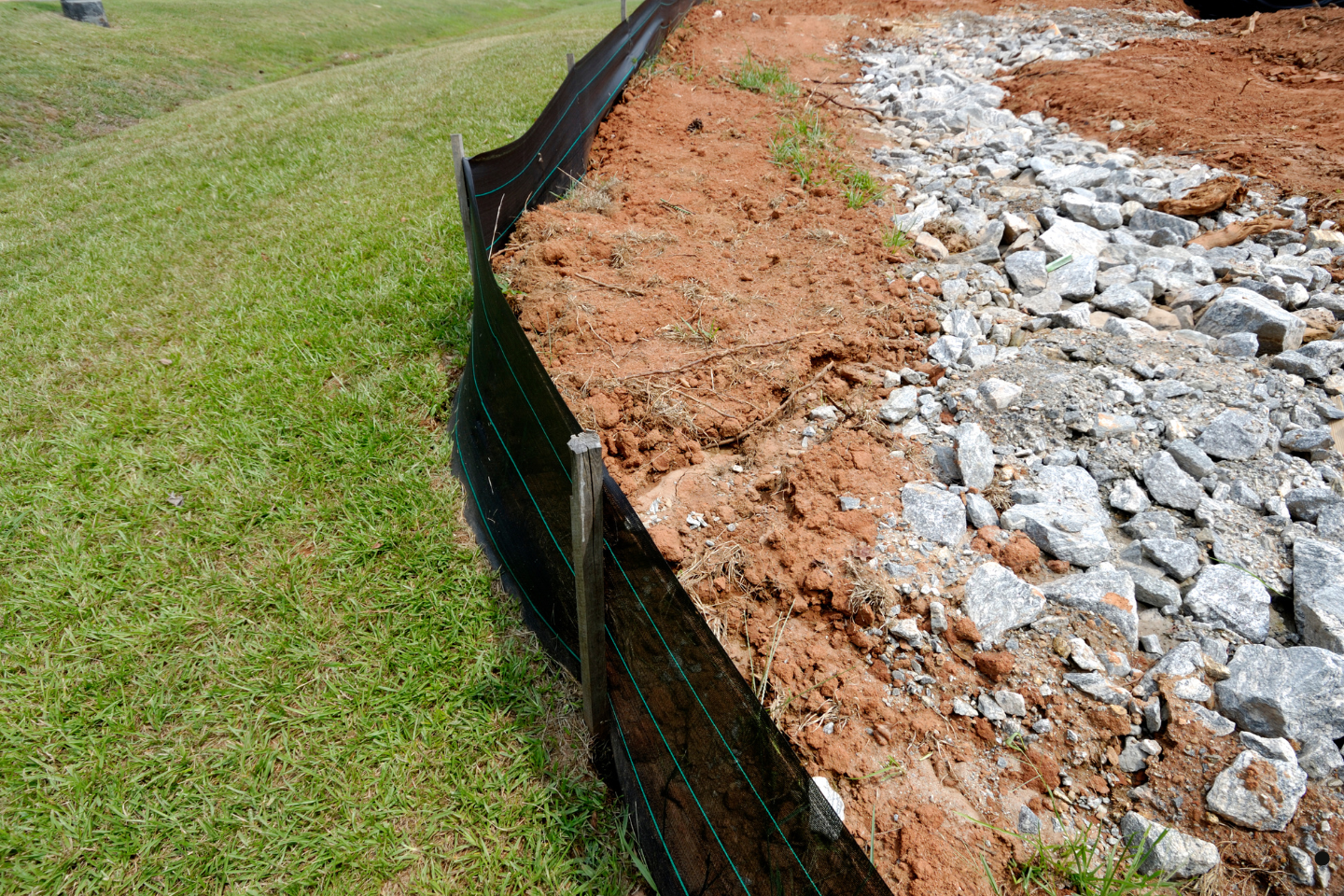 Erosion control silt fence installed along a grassy slope beside exposed soil and rocks.