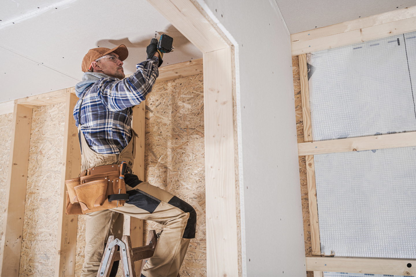 Worker installing drywall while standing on a ladder inside a framed structure.
