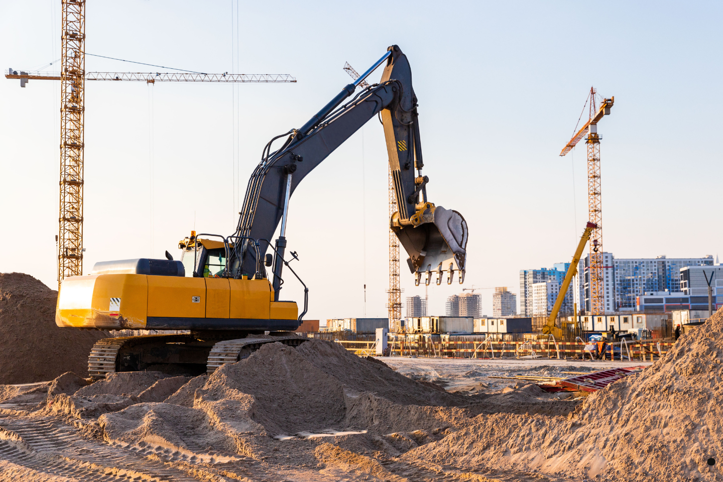 A large excavator digging and moving sand at an active construction site with cranes in the background.