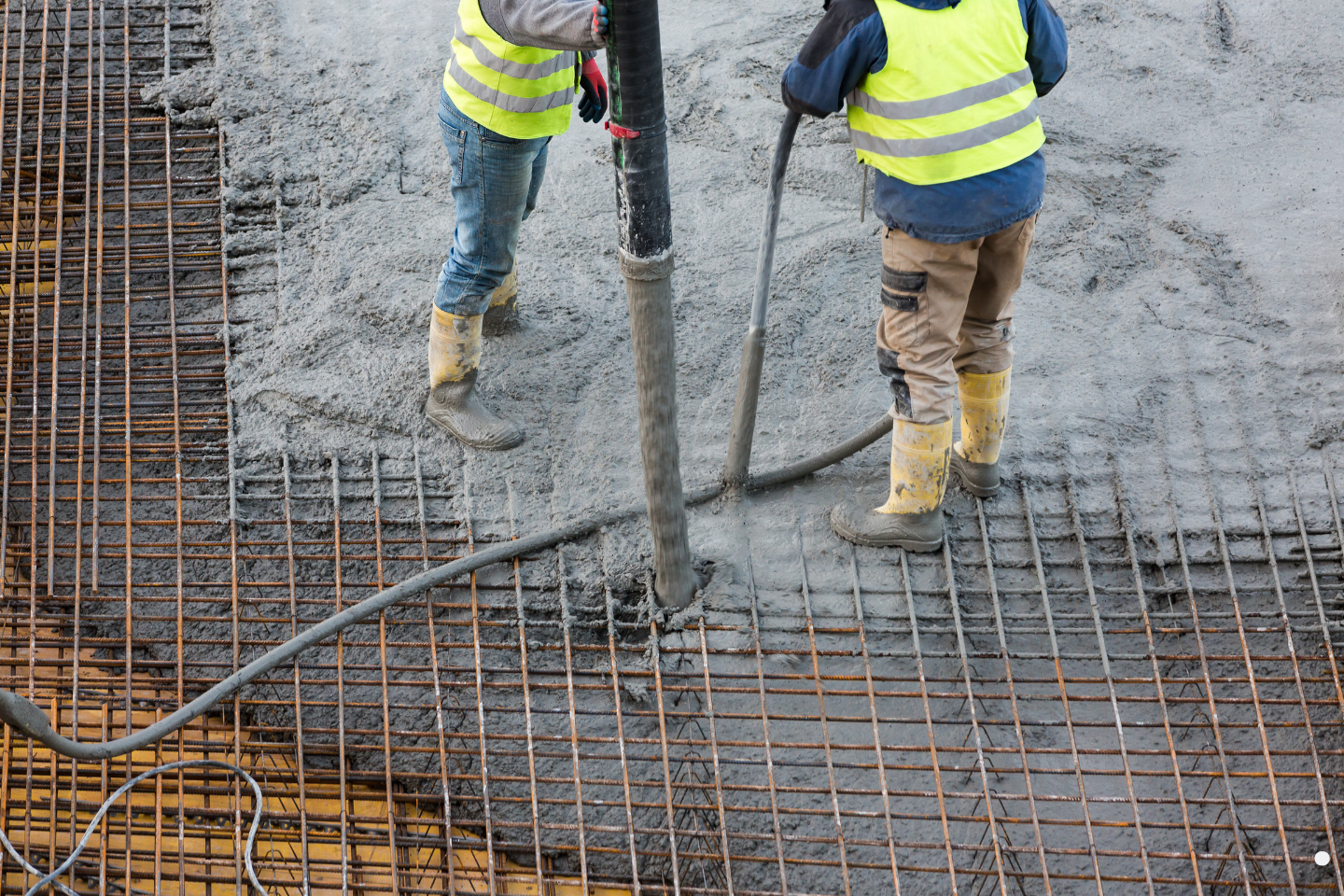 Workers in safety vests pouring concrete over a reinforced rebar grid at a construction site.
