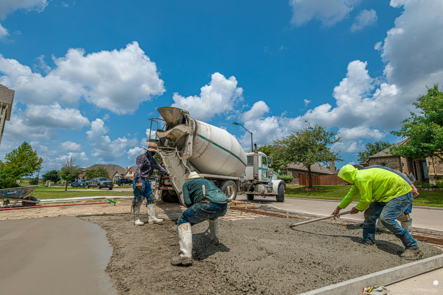 Concrete workers pouring and leveling fresh concrete from a mixer truck in a suburban neighborhood under a bright blue sky.