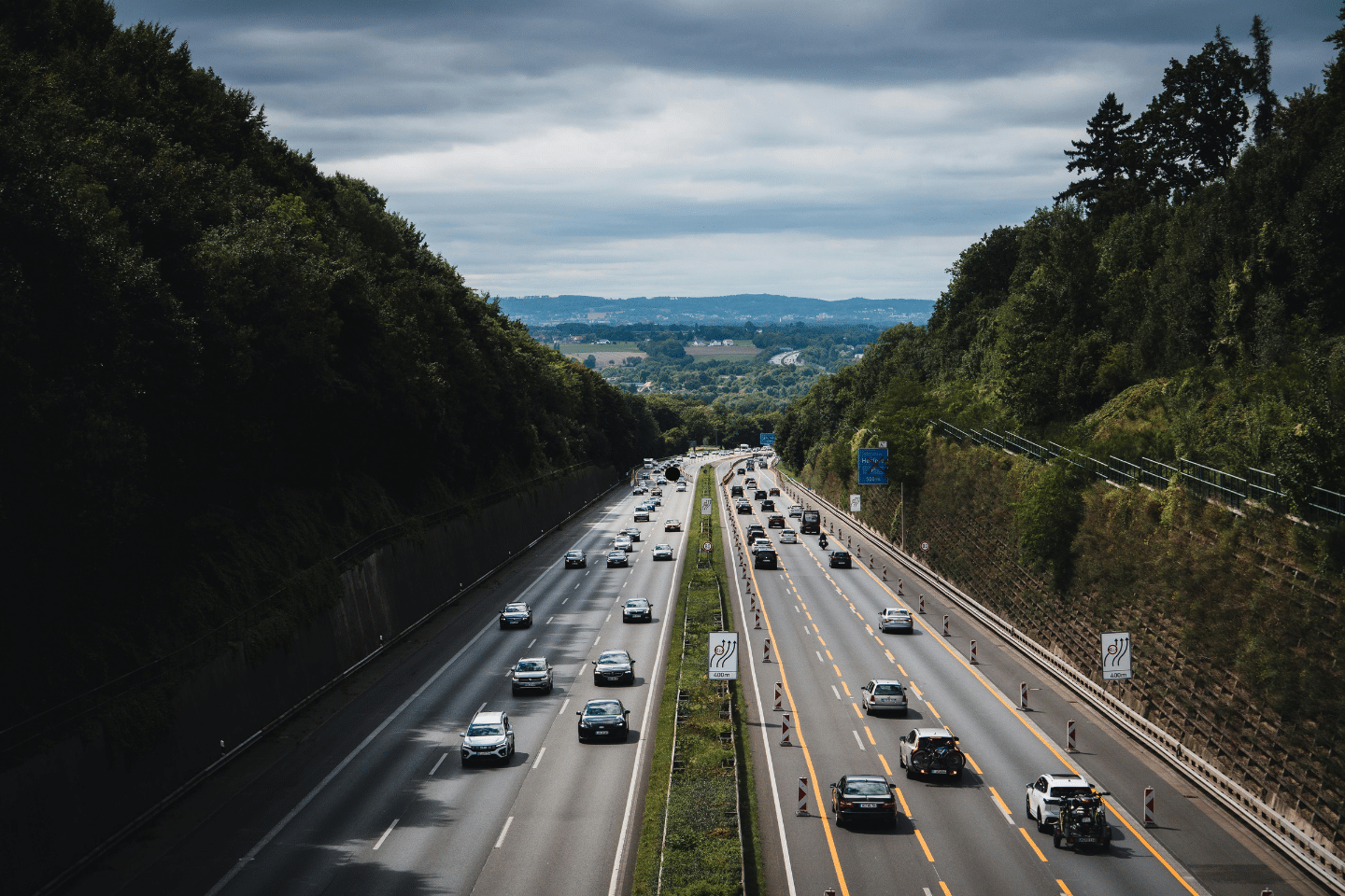 Cars driving along a divided highway surrounded by green trees under a cloudy sky.