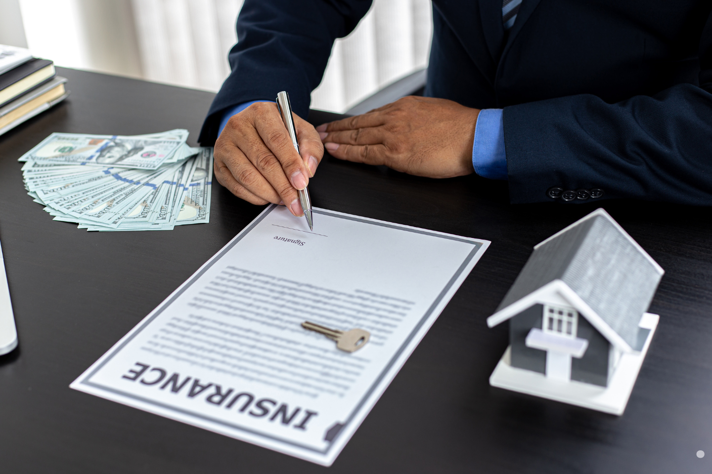 Businessperson signing an insurance contract at a desk with cash and a house model nearby.