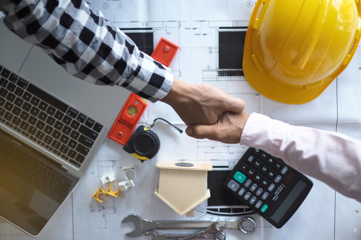 Two people shaking hands over blueprints and construction tools on a worktable.