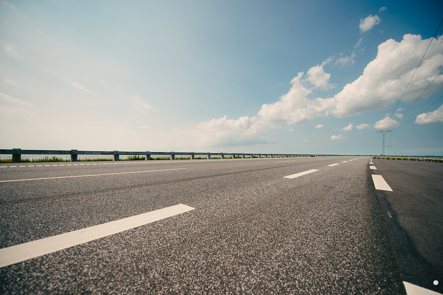 Empty paved highway stretching into the distance beneath a bright sky with scattered clouds.