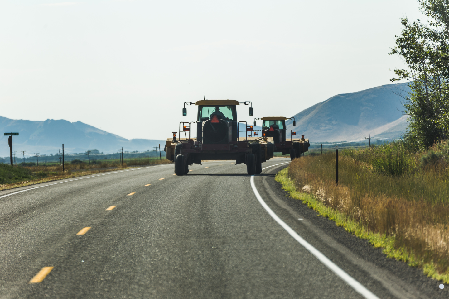 Farm equipment traveling on a rural two-lane road with open fields and distant mountains.