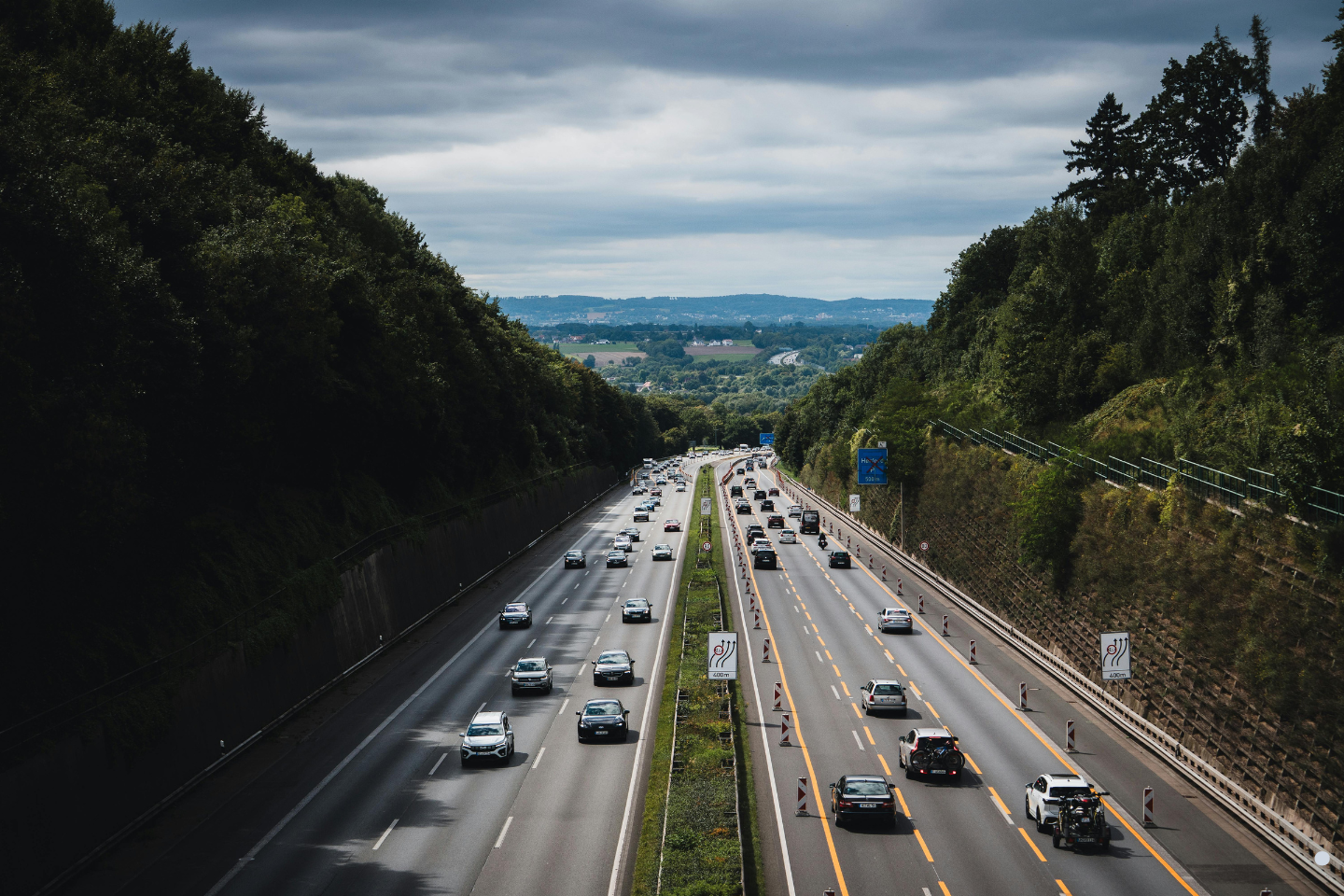 Traffic moving along a divided highway bordered by dense green trees under a cloudy sky.