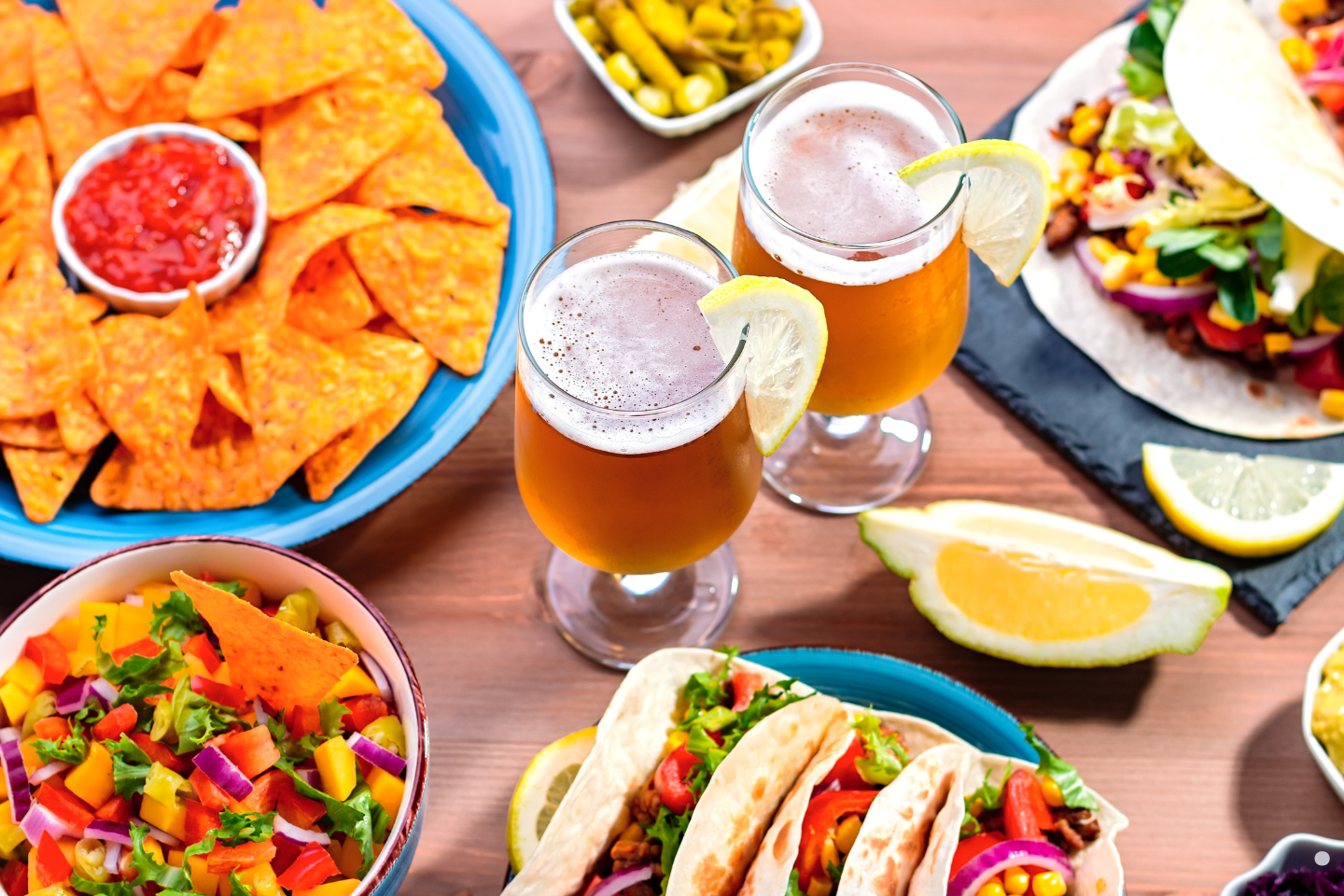 A colorful assortment of tacos, nachos, salsa, and two glasses of beer garnished with lemon.