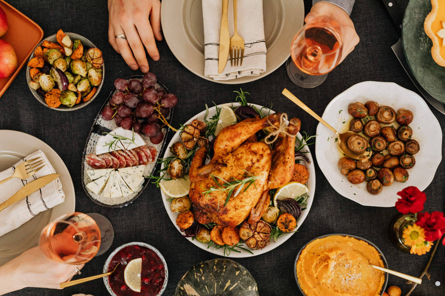 A festive dinner table featuring a roasted chicken, vegetables, grapes, cheese, and glasses of wine.