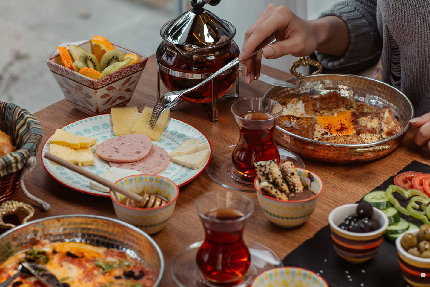 A cozy breakfast spread with cheeses, cold cuts, fruit, tea, and various small dishes on a wooden table.