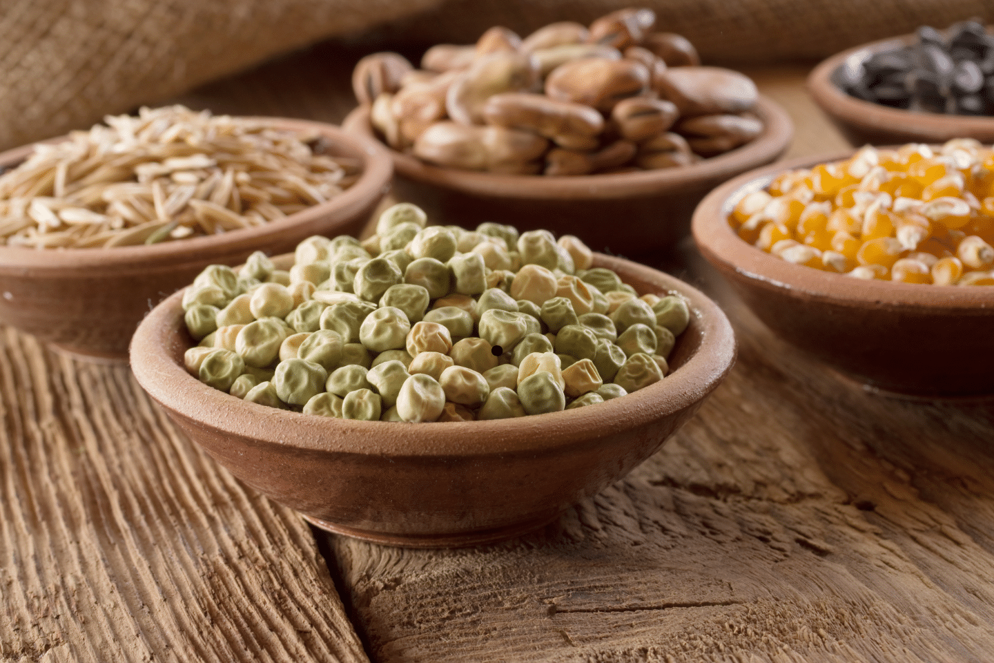 Various dried beans, peas and grains arranged in rustic clay bowls on a wooden surface.