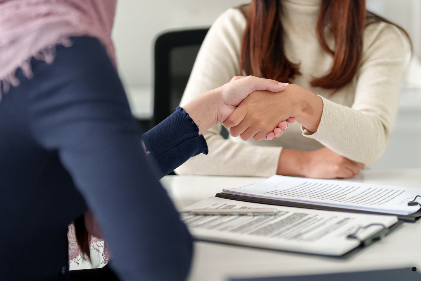 Two people shaking hands across a desk with contracts and paperwork visible.