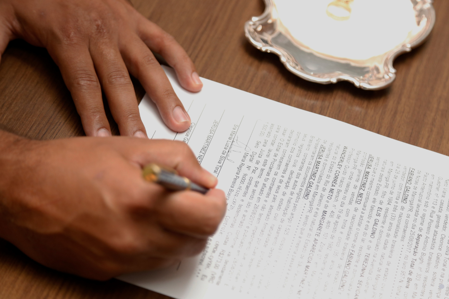 A person signs a legal document on a wooden table with a pen in hand.
