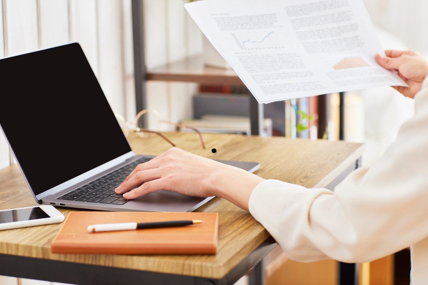 Person working at a laptop while reviewing printed financial documents at a desk.