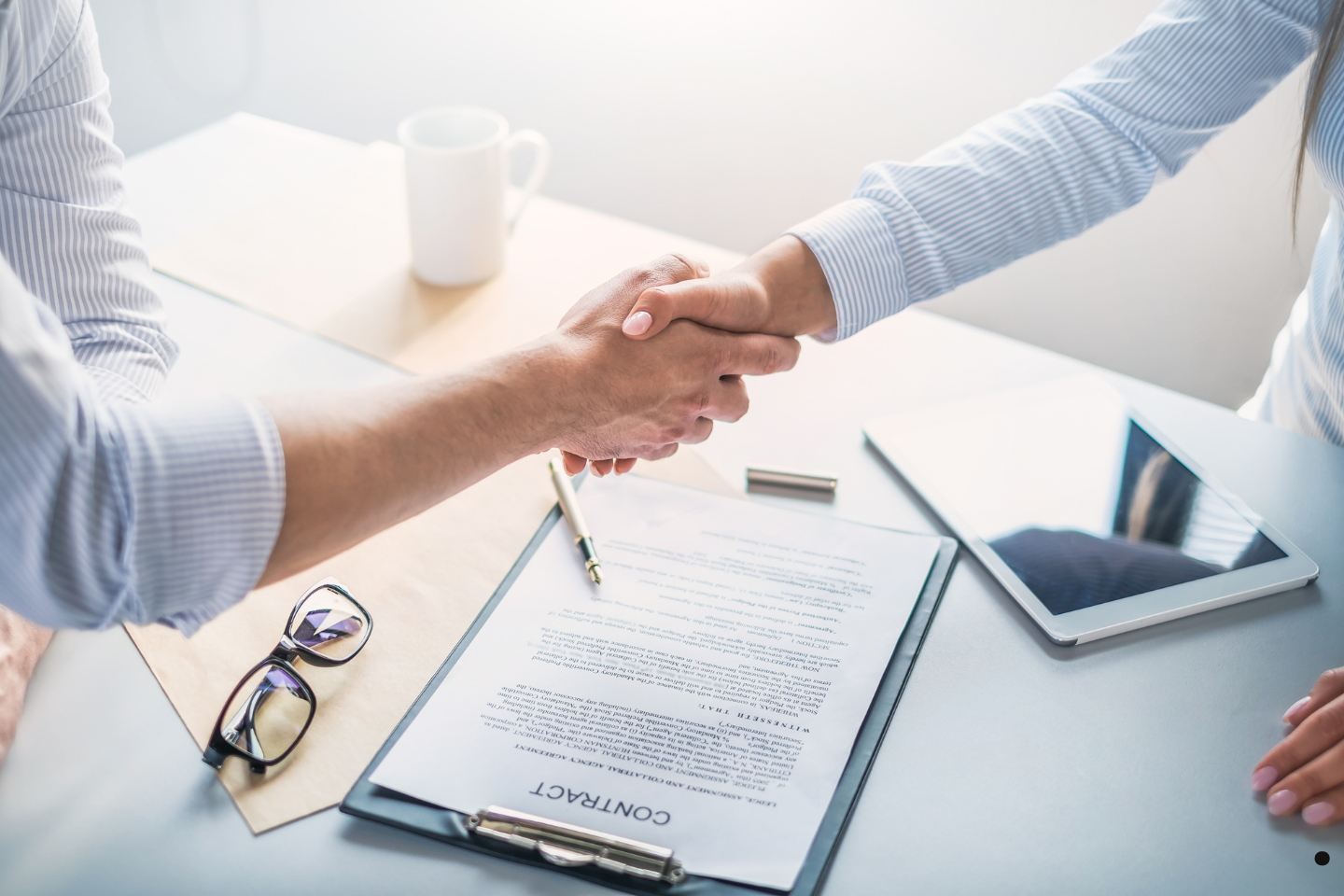 Two people shaking hands over a signed contract on a desk.