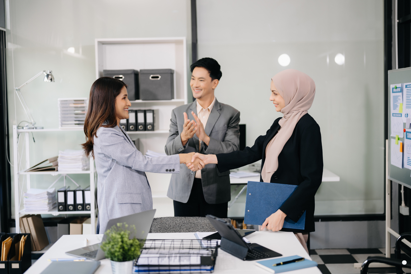 Business professionals smiling and shaking hands while a colleague applauds in an office setting.