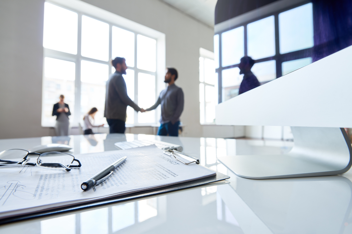 Contract and pen on a desk with two business professionals shaking hands in the background.