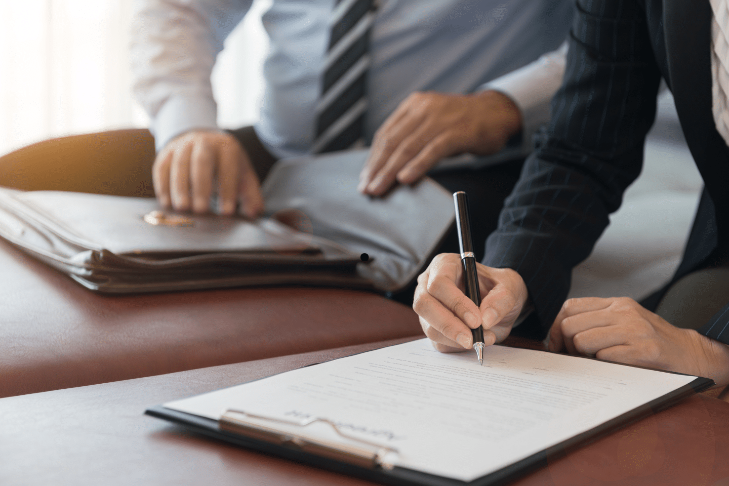 A person signing a formal agreement on a clipboard while another looks on.