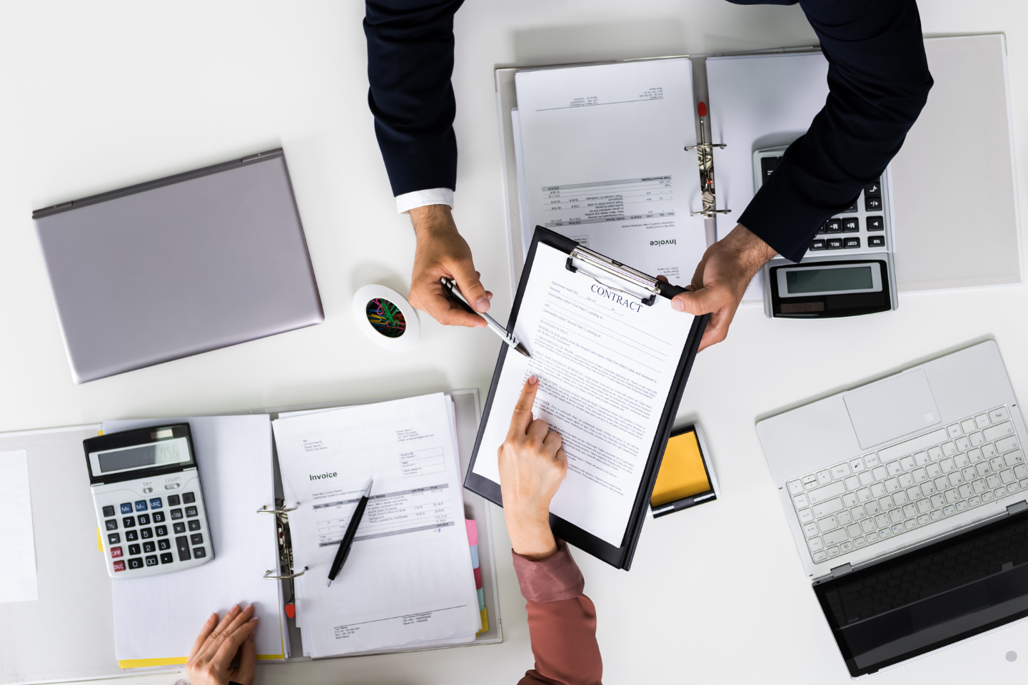 Hands reviewing and signing a contract surrounded by office documents and laptops.
