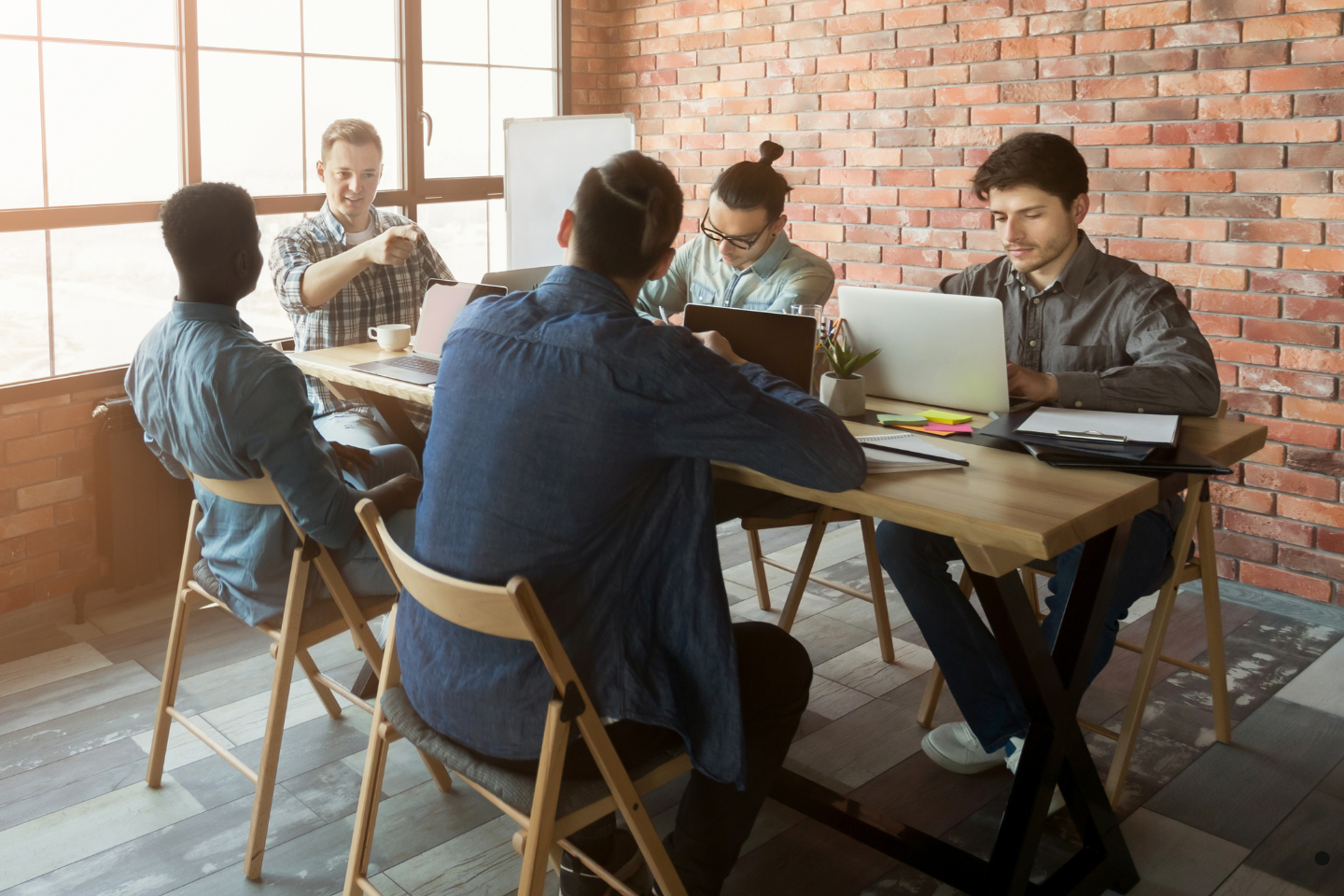 A group of coworkers collaborates at a wooden table in a casual workspace with exposed brick walls.