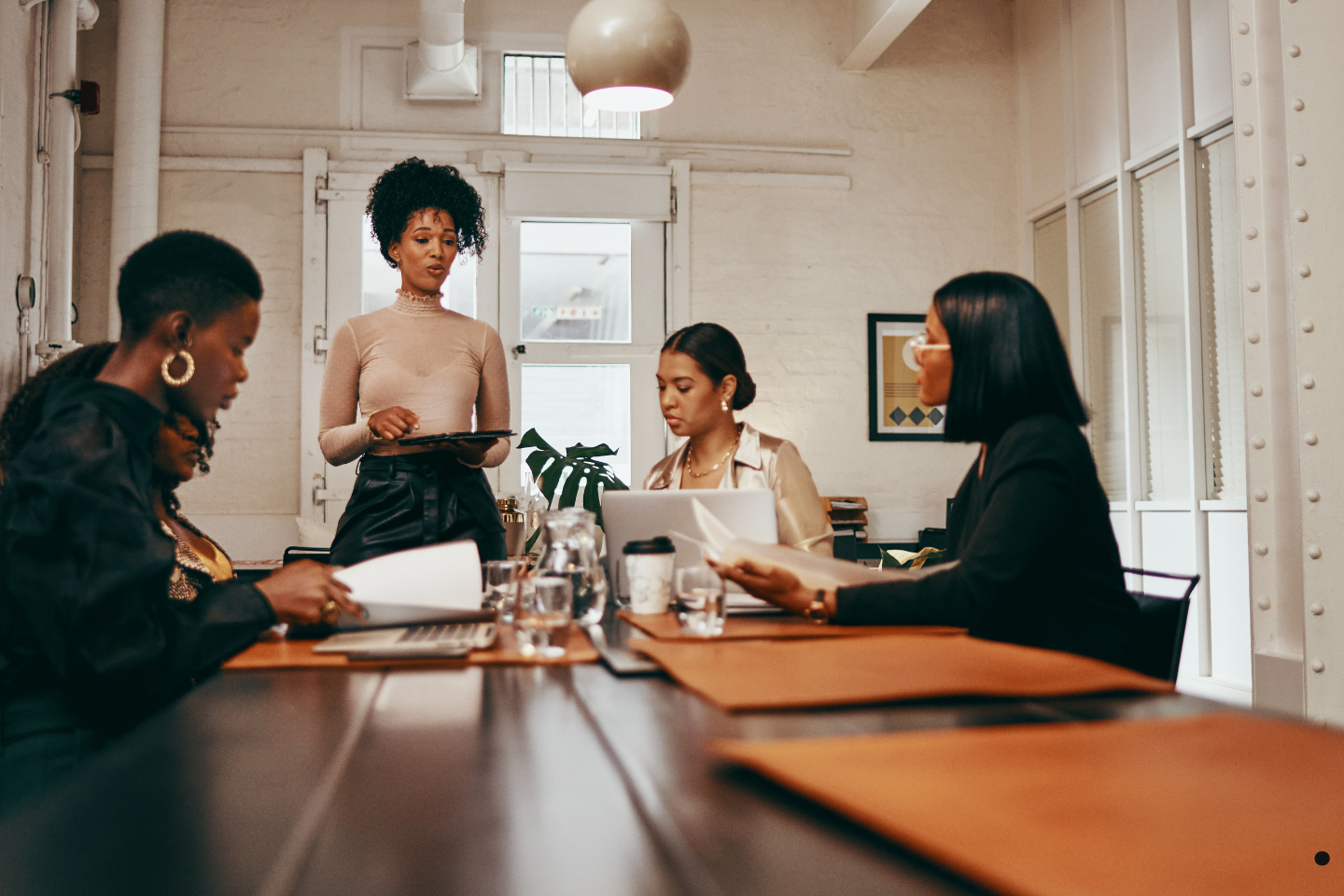 A woman leads a meeting while her team reviews documents and laptops in a warm, collaborative office setting.