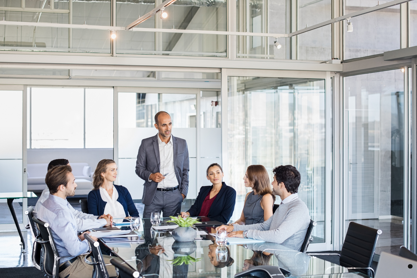 A business leader stands and addresses colleagues seated around a conference table in a modern glass-walled meeting room.