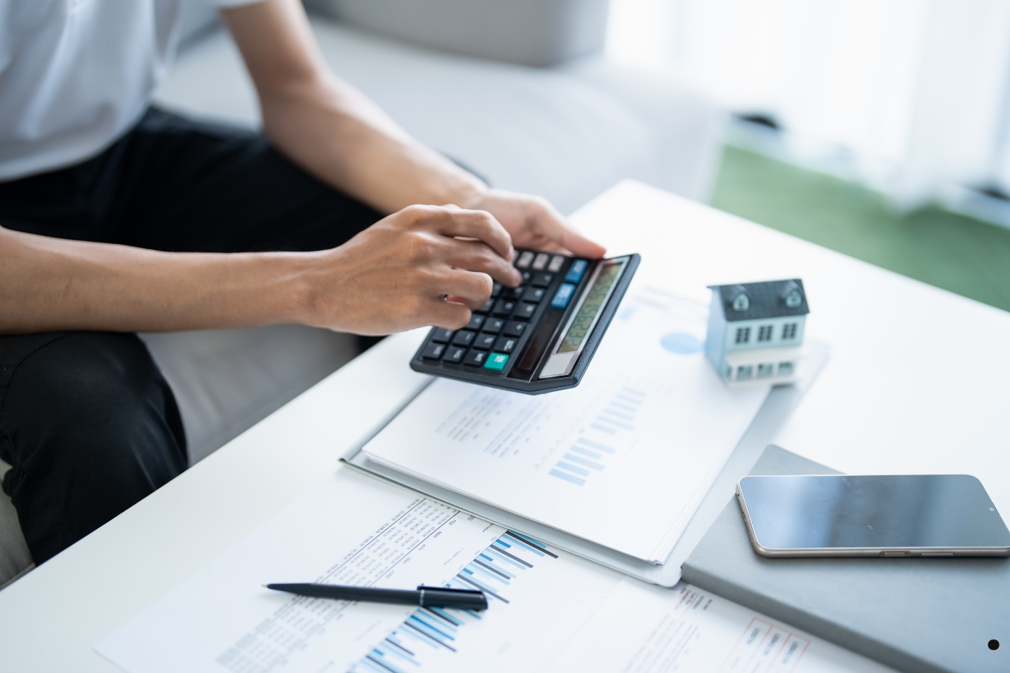 A person calculating finances with documents and a small house model on the table.