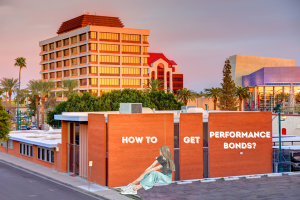 A scenic view of Mesa, Arizona’s urban architecture with a large brick wall showcasing graffiti-style text, 'How to Get Performance Bonds?' in front of a modern building complex and palm trees.