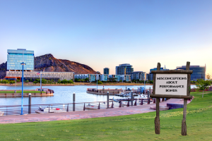 A scenic view of Tempe’s waterfront park with a signboard displaying 'Misconceptions About Performance Bonds,' set against the backdrop of city buildings and a waterfront.