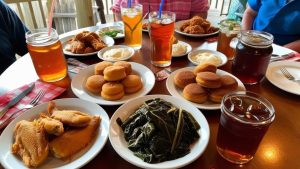 A table spread featuring classic southern dishes in Pine Bluff, Arkansas, including fried catfish, collard greens, cornbread muffins, mashed potatoes, and glasses of sweet tea.