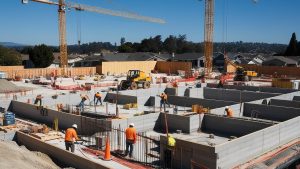 Brentwood, California construction site with workers and heavy machinery building a modern foundation surrounded by residential neighborhoods and cranes in the background.