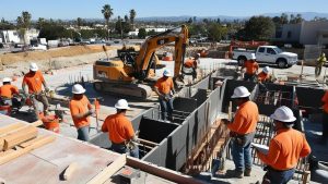 Construction workers in Covina, California, wearing orange safety shirts and helmets, working on a commercial building project with a large excavator in the background.