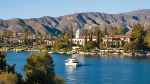 Scenic view of Lake Elsinore, California, with a serene lake, luxurious waterfront homes, palm trees, and the surrounding mountains under a clear blue sky.