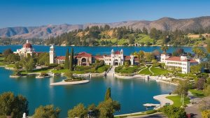 Panoramic view of a picturesque estate and gardens near Lake Elsinore, California, featuring elegant architecture, green landscapes, and the shimmering lake in the background.