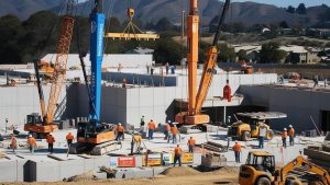 Residential construction project in Morgan Hill, California, with workers and heavy machinery assembling a multi-story building near the hills.