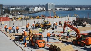 Dynamic construction activity in Newark, California with workers building concrete structures, cranes, and an urban skyline by the waterfront.