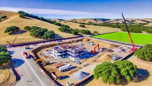 A vibrant construction site in Novato, California, set against rolling hills under a clear blue sky. The site features cranes, trucks, and workers building a steel-framed structure, showcasing the blend of urban development and natural beauty.